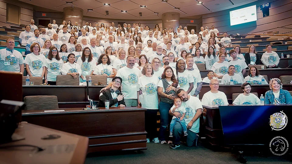 The third Undiagnosed Hackathon, hosted at Mayo Clinic — the whole group gathered on the tiered seats of a lecture hall for a photograph, clinicians, researchers and families together in matching event shirts, with the Undiagnosed Hackathon and Wilhelm Foundation mark in the lower corner