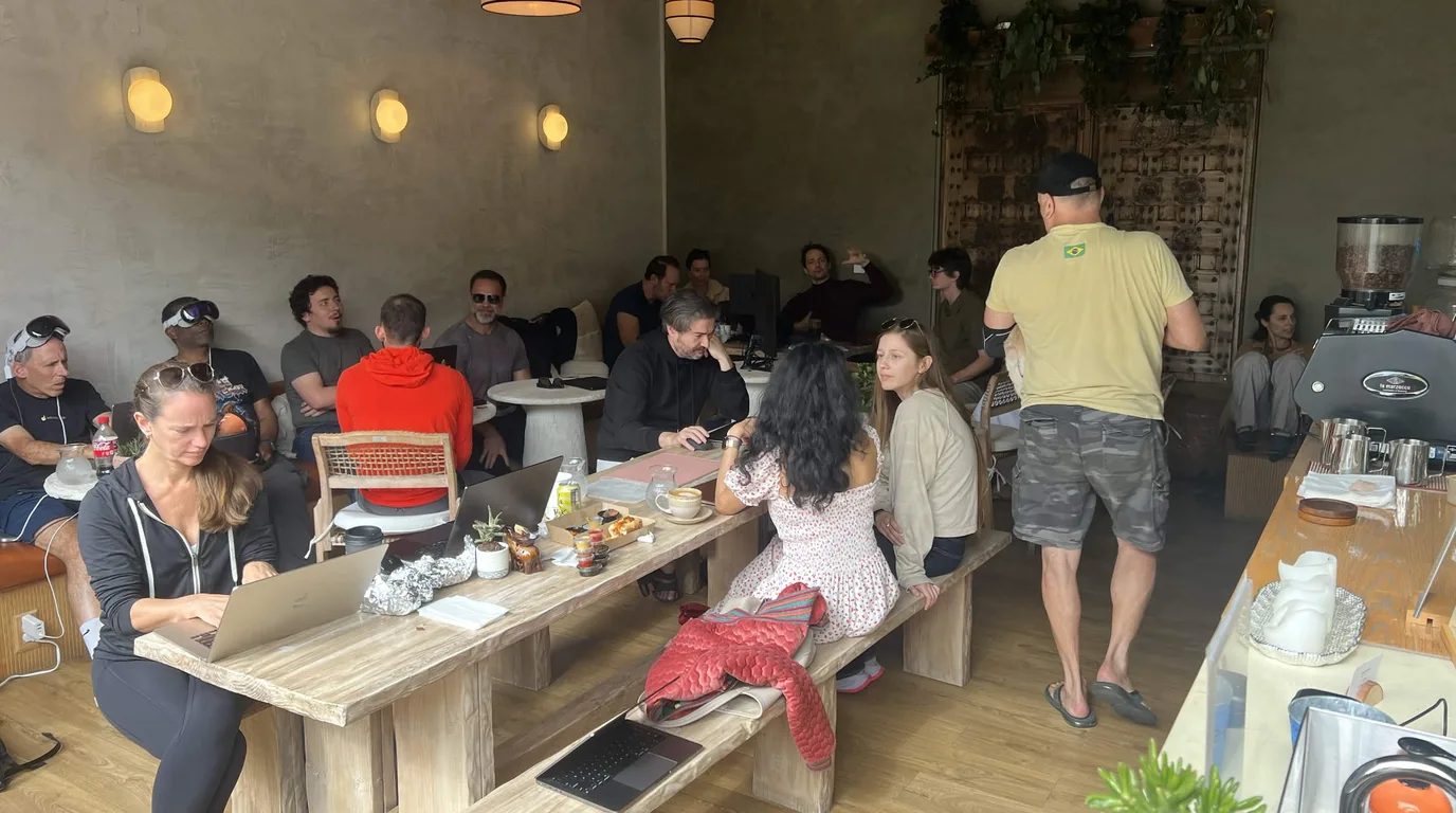 Photograph inside a Venice cafe on a Science Sunday — people filling a long communal wooden table with laptops, coffee and pastries, more seated around low tables behind, an espresso machine along the right wall